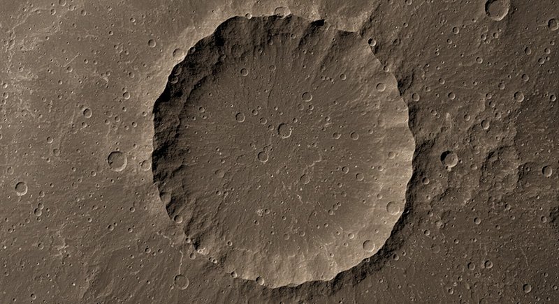 Ein großer, kreisförmiger Einschlagskrater mit steilen Wänden und einem rauen Boden ist auf der felsigen, kraterübersäten Oberfläche des kleinsten Planeten zu sehen.