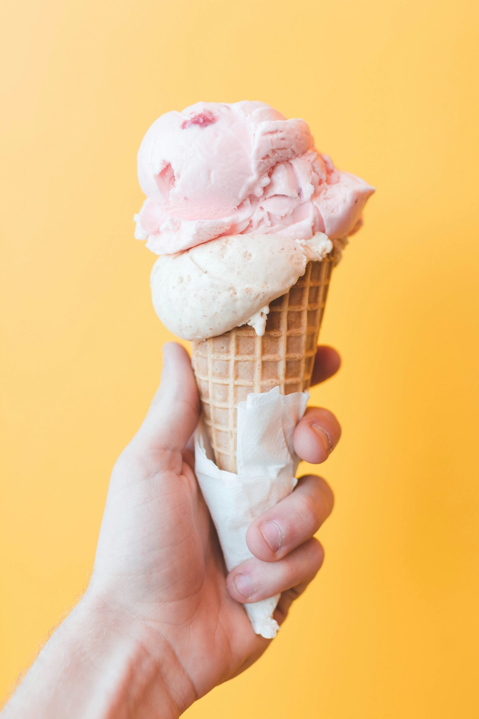 Hand holding a double scoop of strawberry ice cream in a waffle cone against a yellow background.