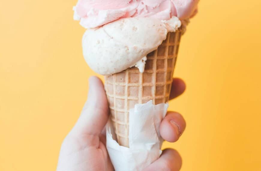 Hand holding a double scoop of strawberry ice cream in a waffle cone against a yellow background.