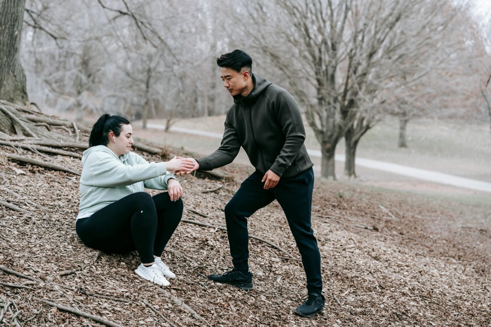 A man assists a woman as she rises from the ground during an outdoor workout in a wooded area.