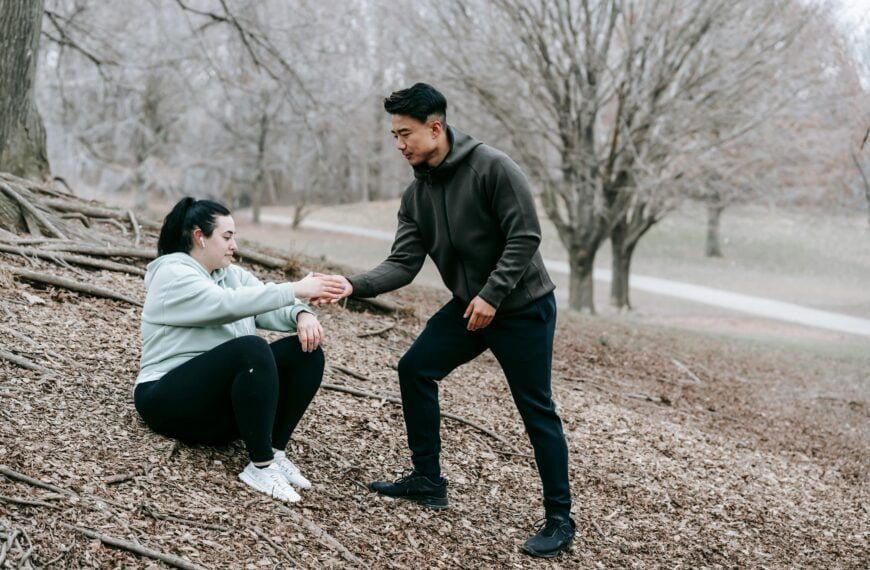 A man assists a woman as she rises from the ground during an outdoor workout in a wooded area.