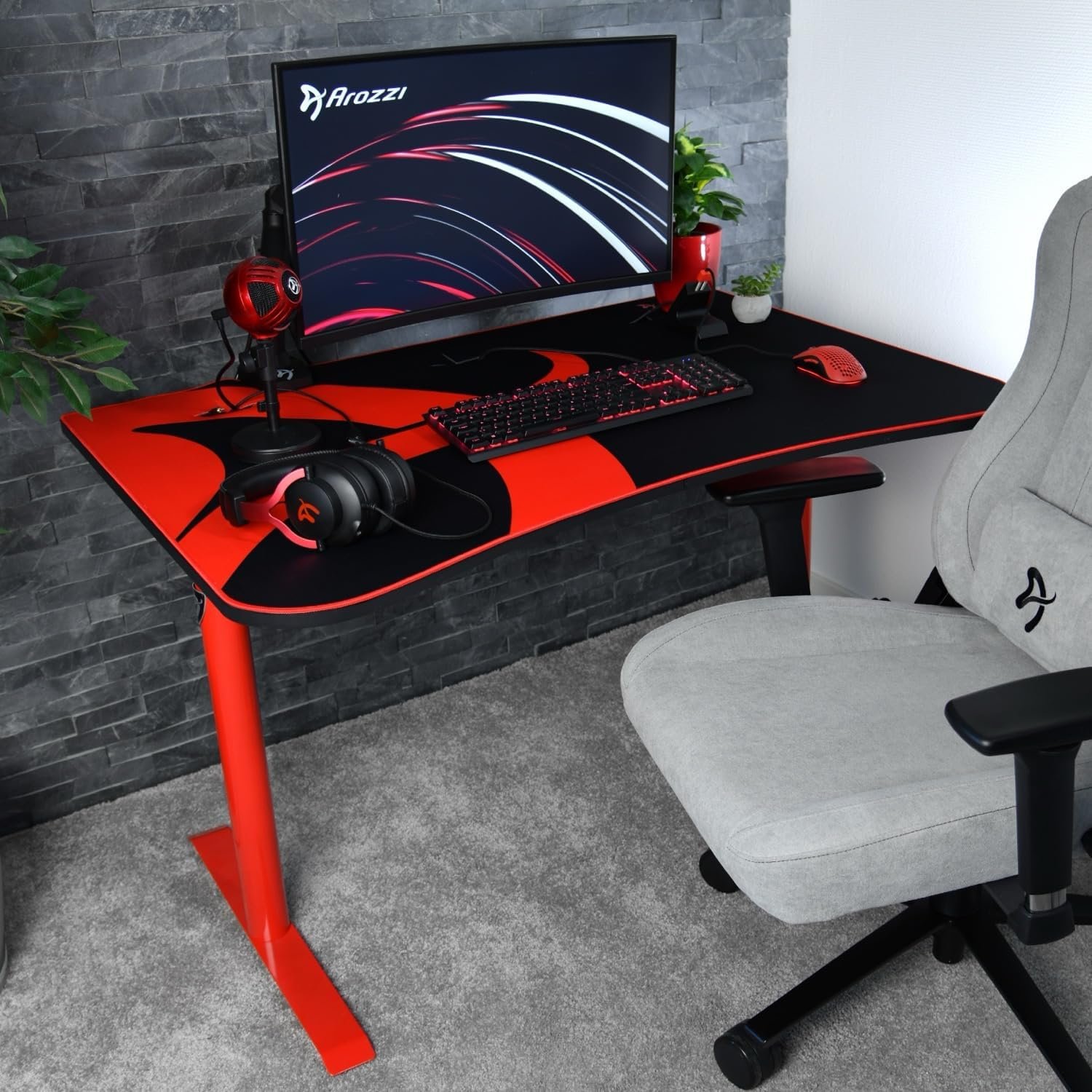 A computer desk setup with a monitor, keyboard, mouse, and headphones, all featuring a black and red color scheme. A grey chair is positioned at the desk, which is placed against a textured wall.