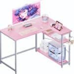A pink and gray desk setup includes a monitor displaying an animated character, keyboard, mouse, gaming console, tablet, and various accessories including a headphone stand and desktop computer.