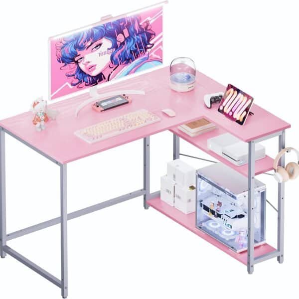 A pink and gray desk setup includes a monitor displaying an animated character, keyboard, mouse, gaming console, tablet, and various accessories including a headphone stand and desktop computer.