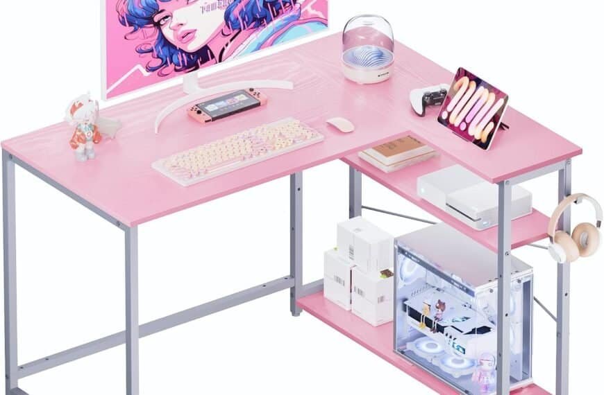 A pink and gray desk setup includes a monitor displaying an animated character, keyboard, mouse, gaming console, tablet, and various accessories including a headphone stand and desktop computer.