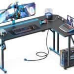 A modern gaming desk setup featuring a wide monitor displaying a game, a keyboard, mouse, gaming console, laptop, and accessories like headphones and a small lamp, with LED lighting under the desk.