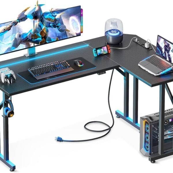 A modern gaming desk setup featuring a wide monitor displaying a game, a keyboard, mouse, gaming console, laptop, and accessories like headphones and a small lamp, with LED lighting under the desk.