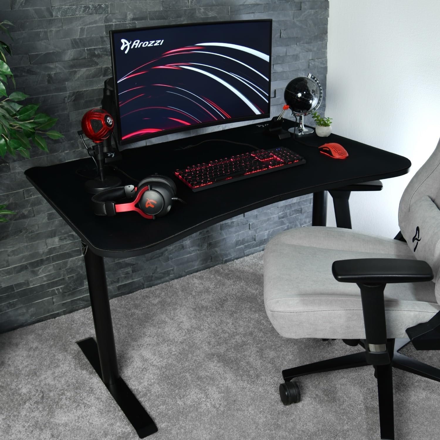 A gaming setup with a monitor, keyboard, mouse, speakers, and a headset on a black desk. There is a gray chair in front of the desk and a small plant in the corner.