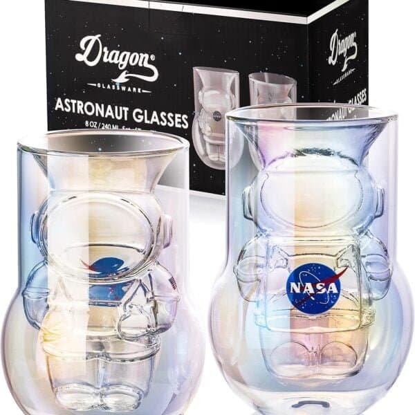 Two astronaut-themed glasses are displayed in front of their packaging. The glasses feature an astronaut-shaped design inside and have NASA and a spaceship logos. The packaging is branded "Dragons Glassware.