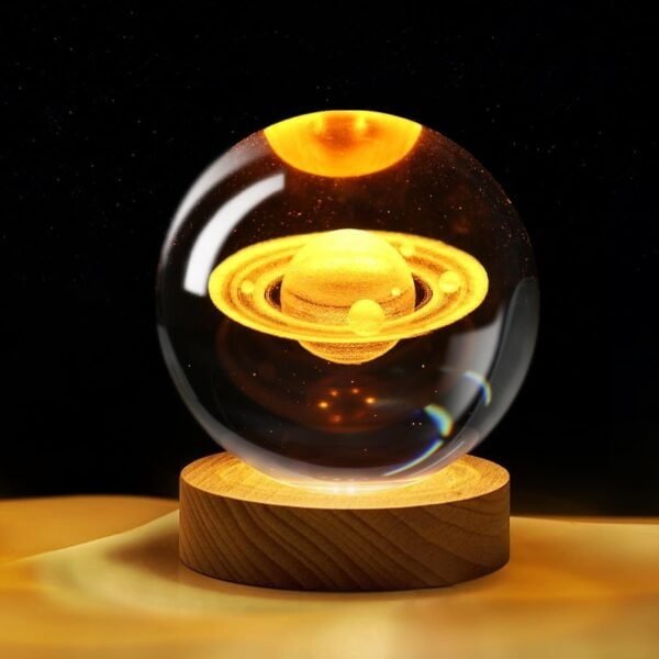 A glass orb displays a glowing, miniature model of Saturn with rings, resting on a wooden base against a dark background.