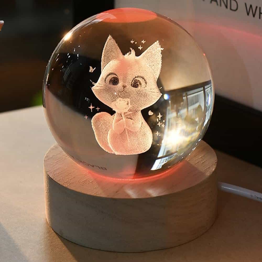A glass orb on a wooden base with a 3D etched image of a cute, cartoon-like fox inside, illuminated by a warm light.