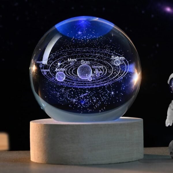 A crystal sphere displaying an engraved solar system model, with labeled planets orbiting the Sun, placed on a wooden stand. An astronaut figurine is partially visible on the right.