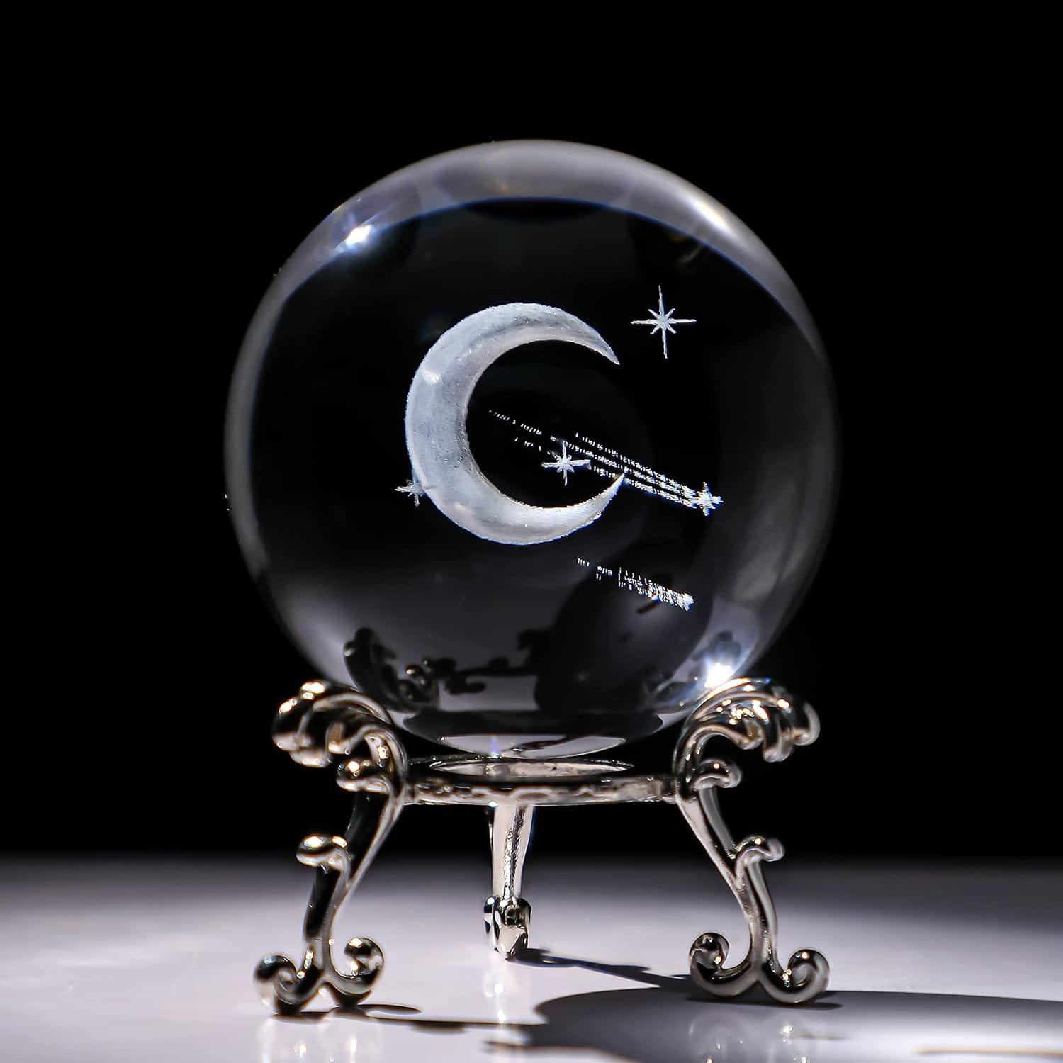 A glass orb with a crescent moon and stars etched inside, placed on an ornate metallic stand against a dark background.