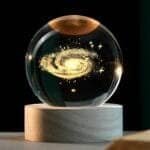 Glass orb with a galaxy design inside, resting on a wooden base against a dark background.