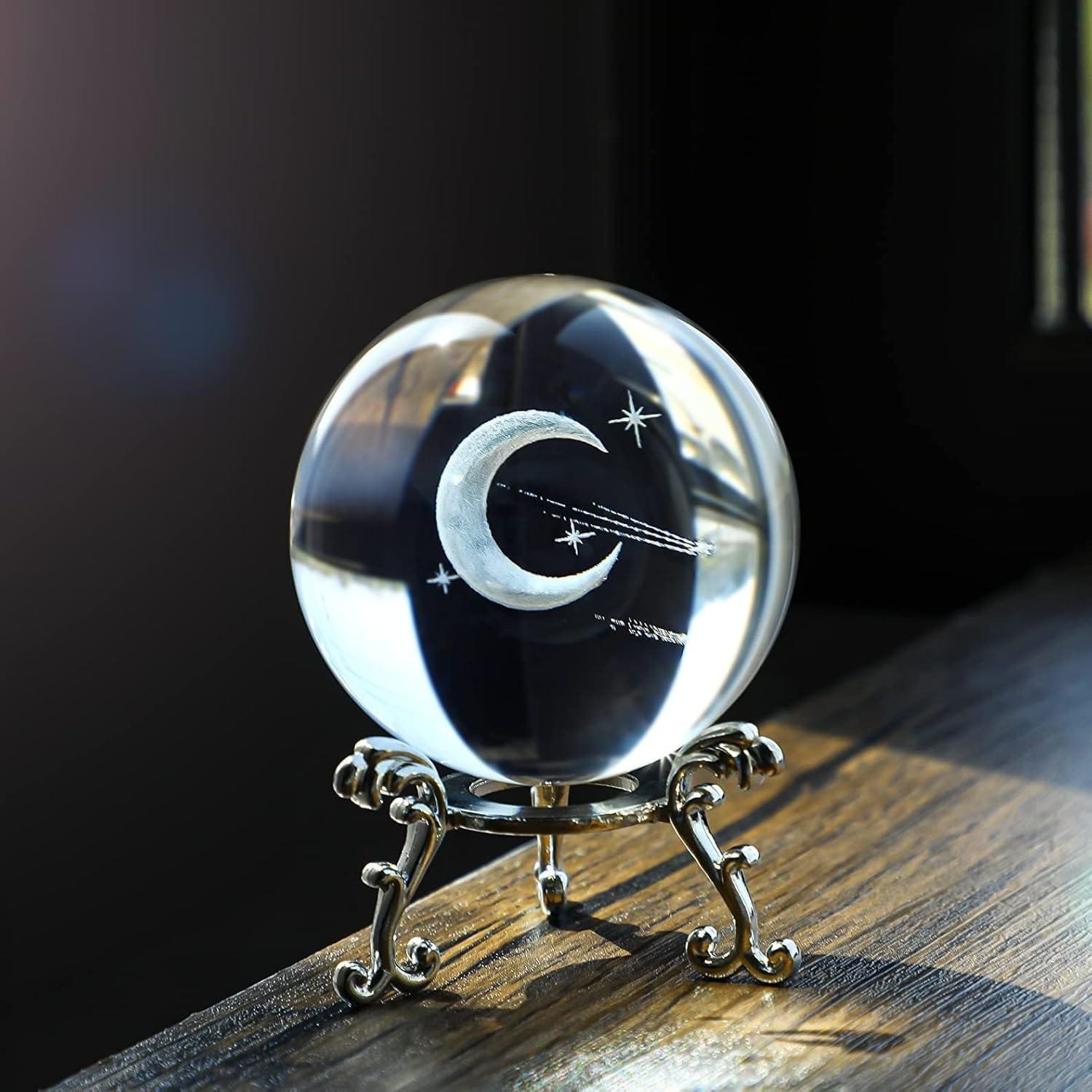 Glass crystal ball with an etched crescent moon and stars design, sitting on an ornate silver stand on a wooden surface.