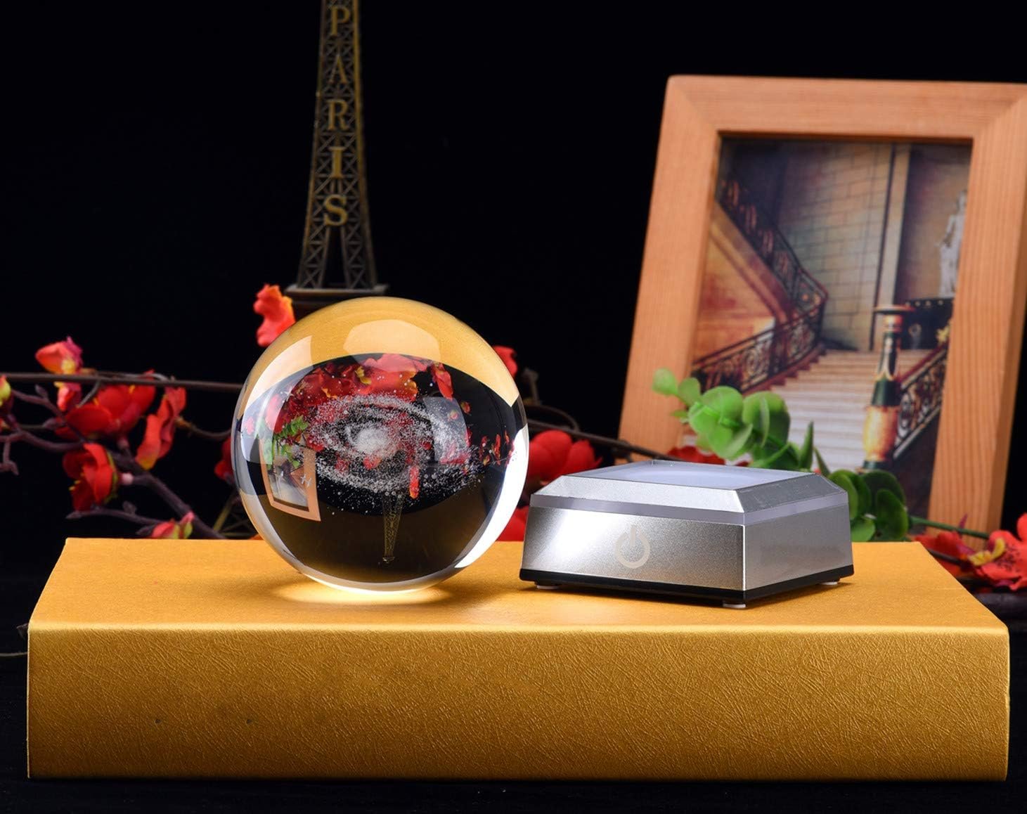 A crystal ball with a galaxy pattern sits on a gold box next to a silver square device. In the background are red flowers, a framed picture, and a mini Eiffel Tower statue.
