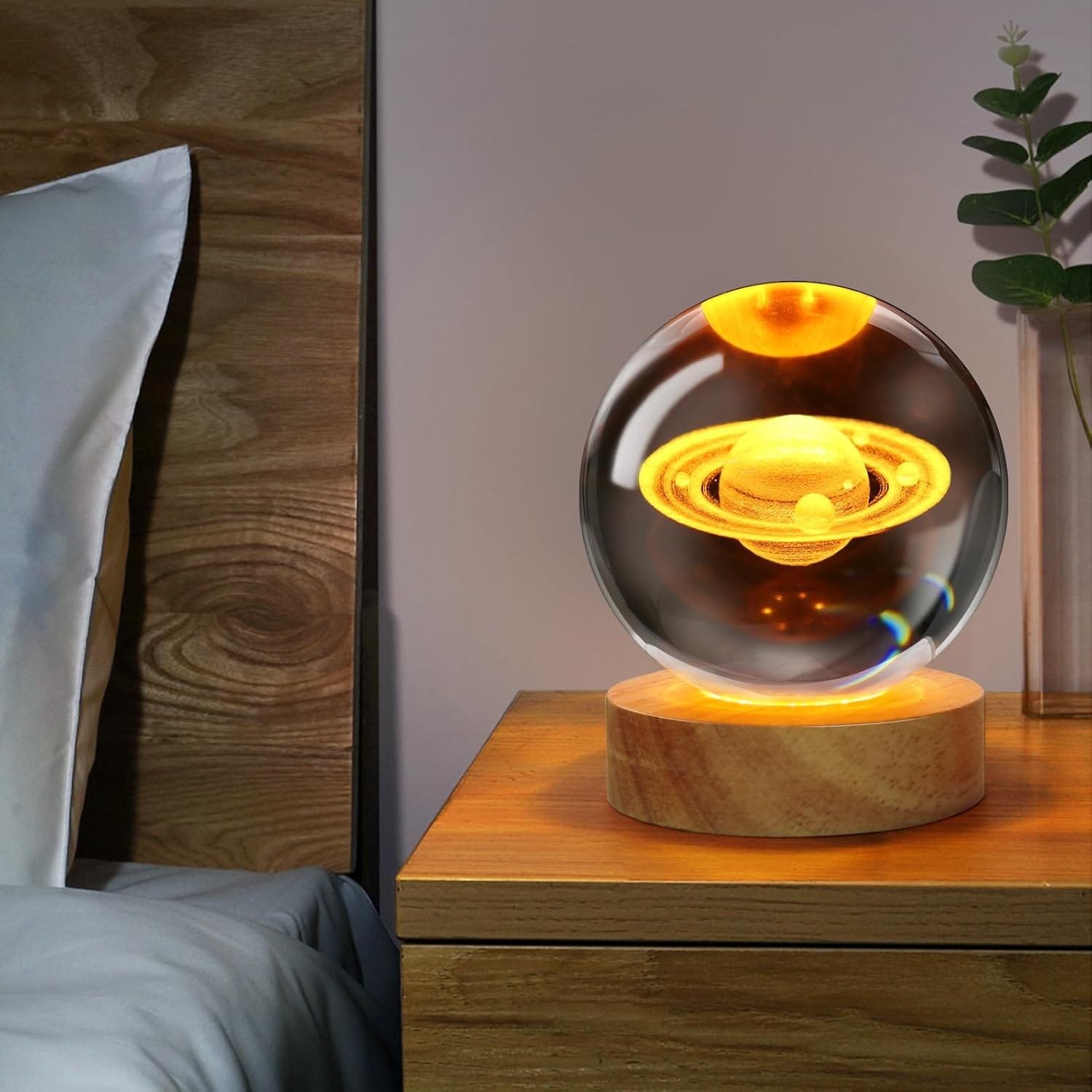 A glowing planet-themed glass orb on a wooden stand, placed on a bedside table next to a plant.