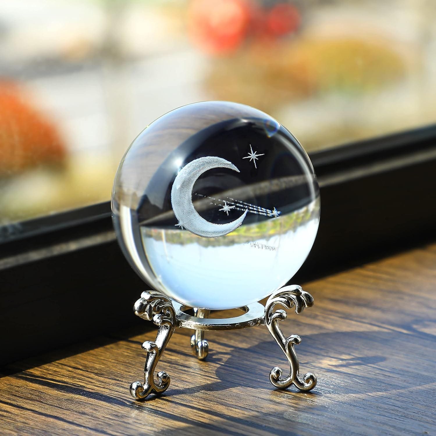 A glass orb with a crescent moon and stars design, resting on an ornate silver stand on a wooden surface near a window.