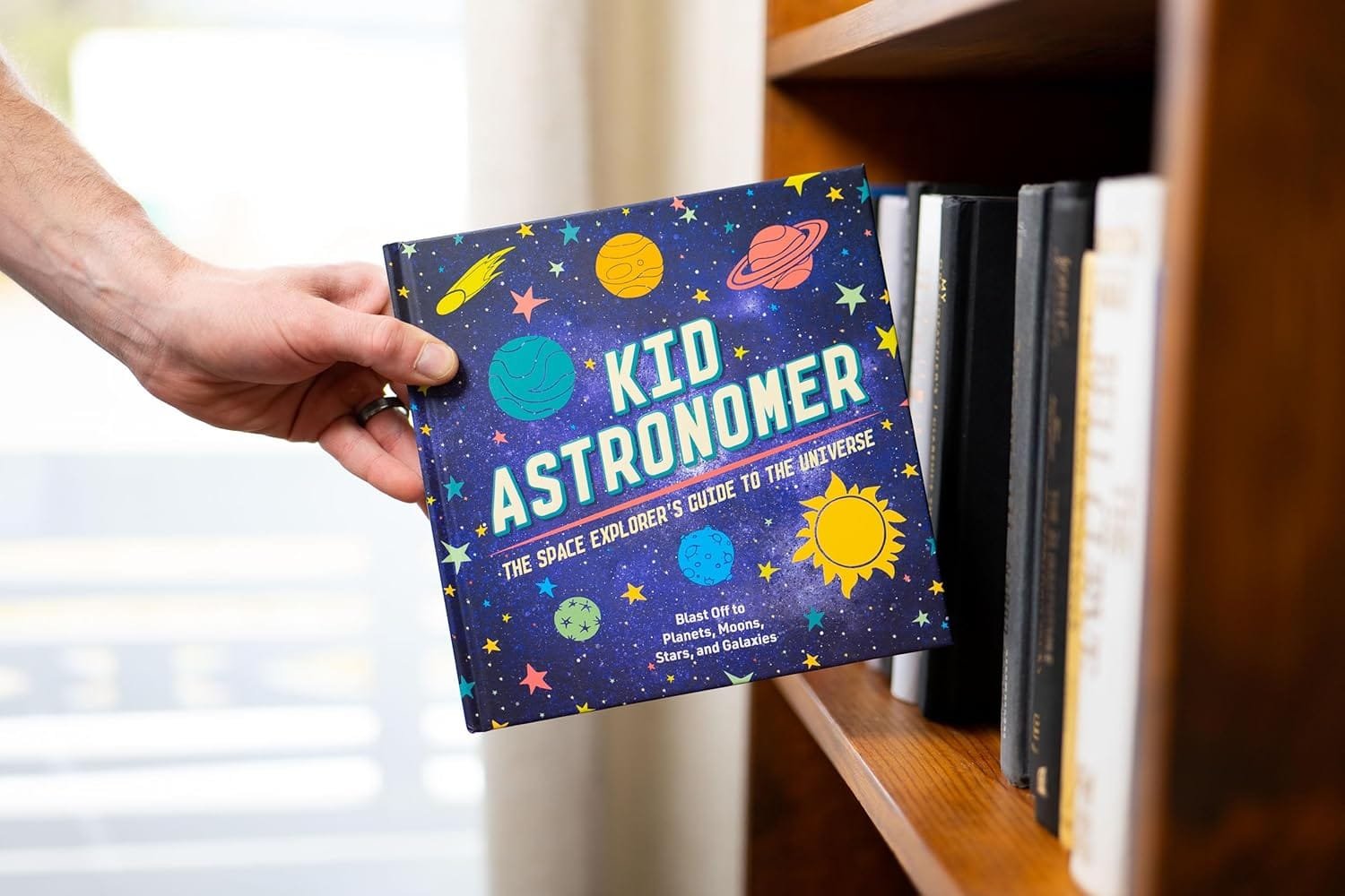 A hand holds a book titled "Kid Astronomer: The Space Explorer's Guide to the Universe" from a bookshelf.