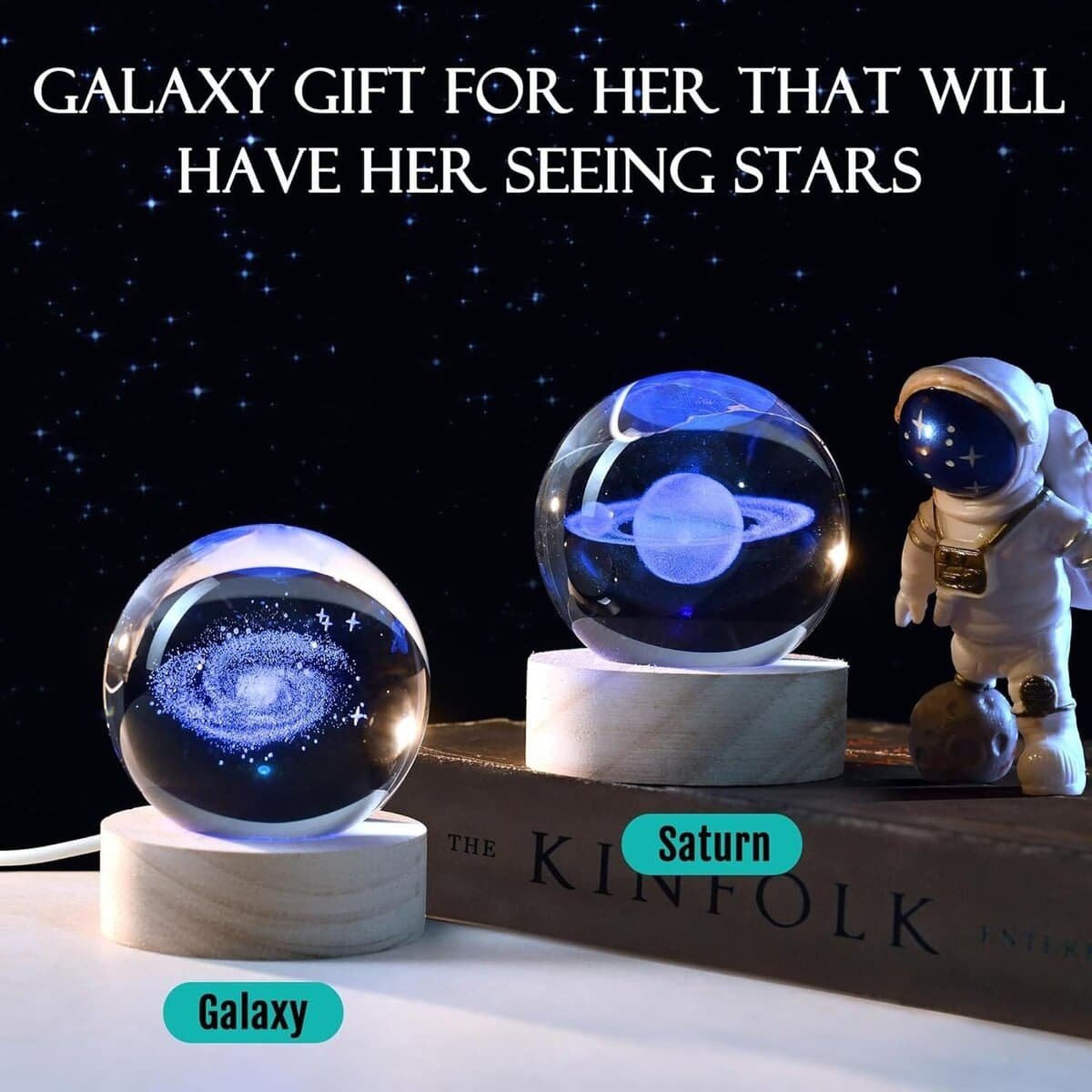 Crystal globes with galaxy and Saturn designs are displayed on a wooden base next to a small astronaut figure and a book. Text above reads: "Galaxy gift for her that will have her seeing stars.