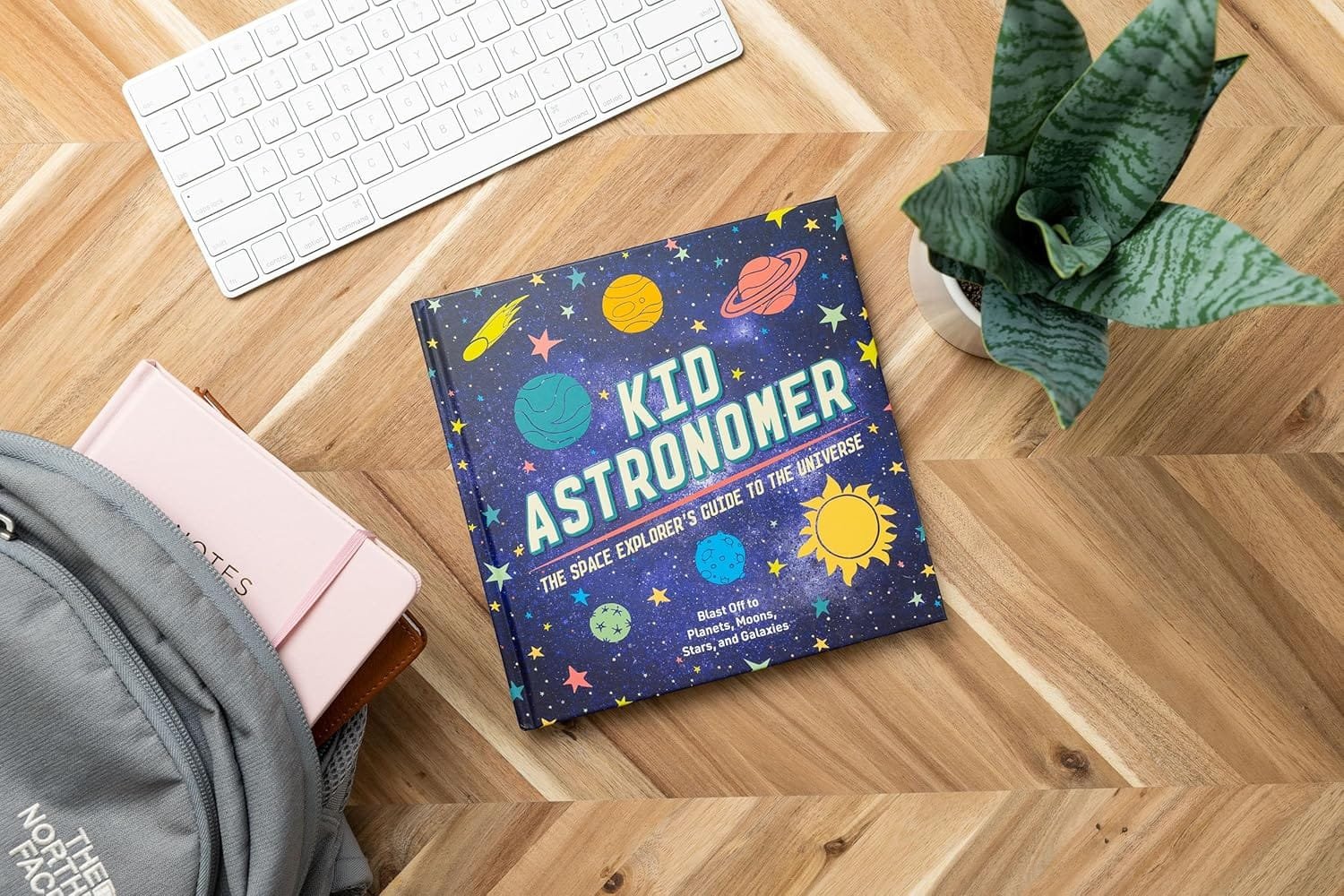 A children's book titled "Kid Astronomer" lies on a table next to a keyboard, a plant, and a backpack.