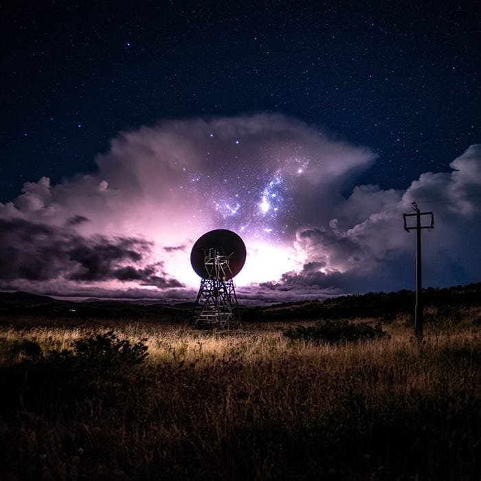 A large satellite dish silhouette stands against a starry night sky, where illuminated clouds hover above the grassy terrain, capturing the mysterious beauty of gamma-ray emissions.