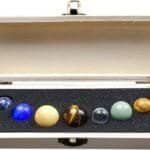 A wooden box containing eight marbles of various colors and materials, arranged in a row on black foam padding.