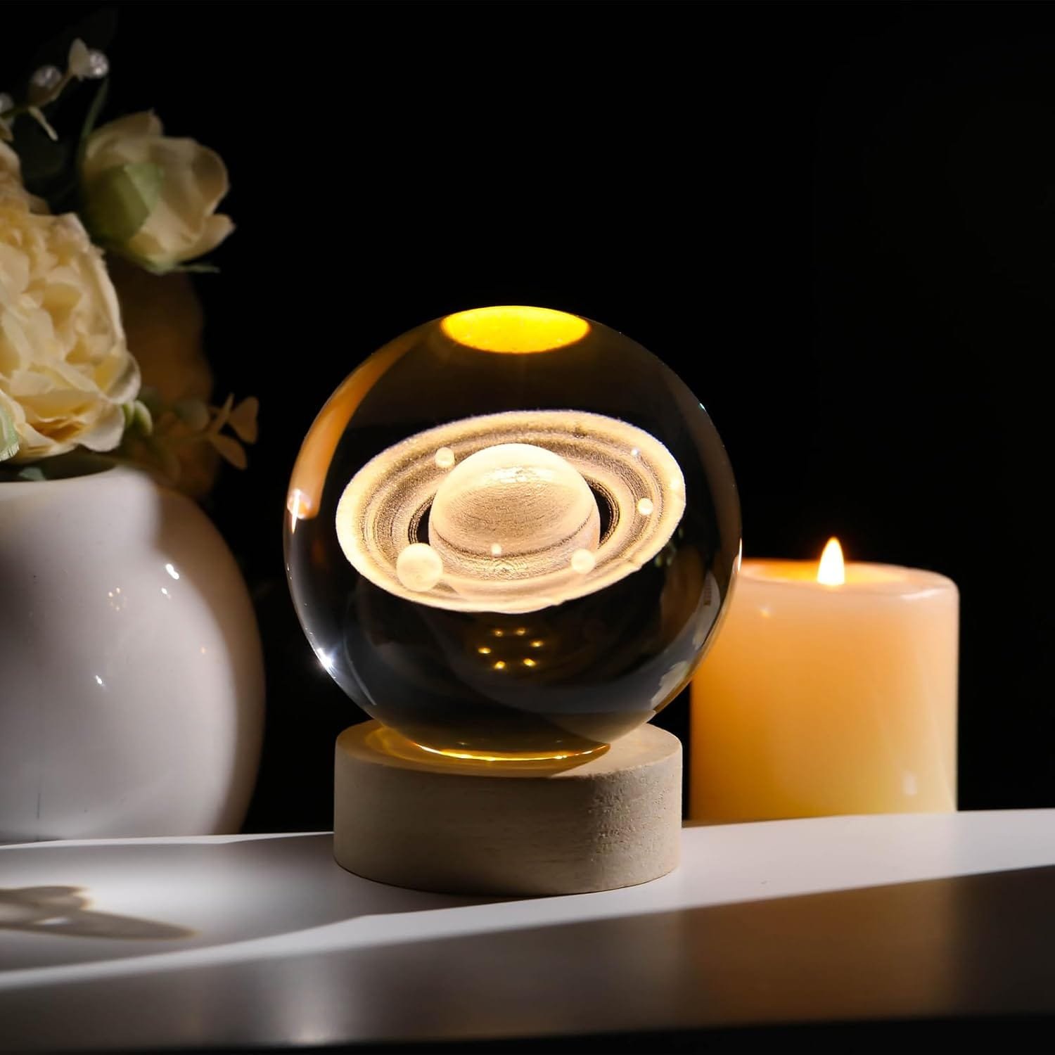 Glass orb with etched Saturn design on a wooden base, positioned next to a lit candle and a vase with white flowers.