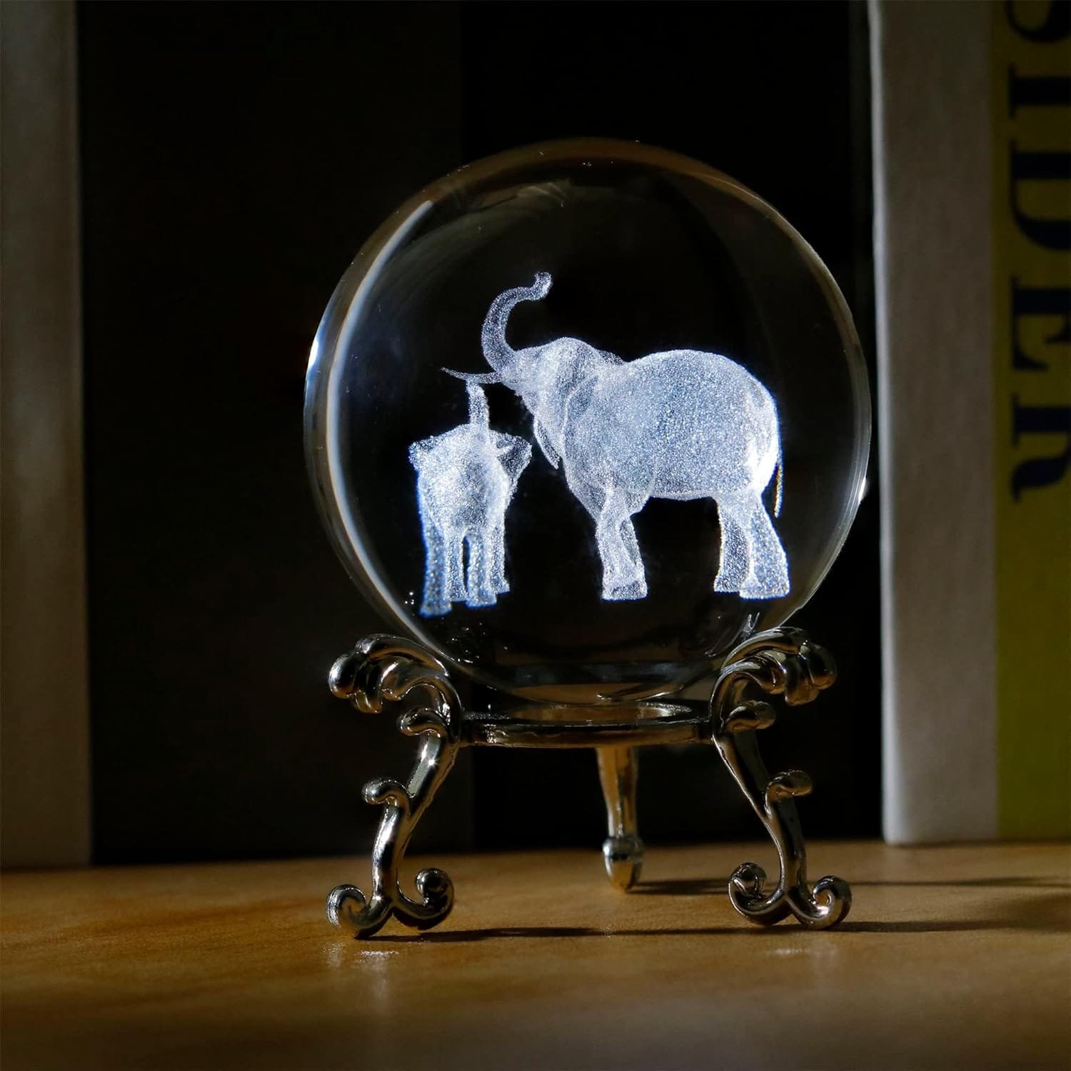 A glass sphere on a stand displays a luminescent sculpture of two elephants, one larger with a raised trunk and one smaller, set against a dark background.