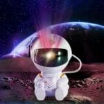 A small astronaut figurine sits on a moon-like surface with a glowing helmet, all brought to life by the Your World with the Astronaut Galaxy Projector. A planet and stars shimmer in the background, creating a mesmerizing cosmic scene.