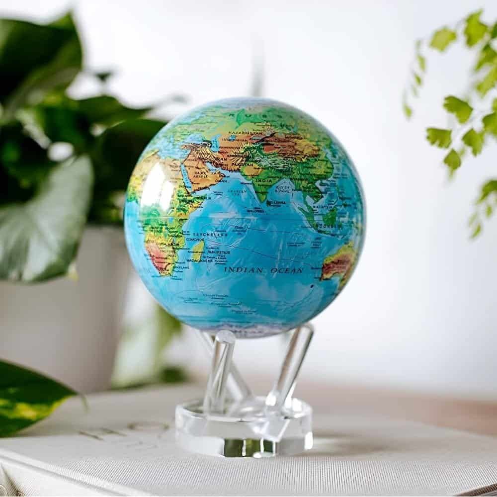 The Globe Relief Map Blue 6" on clear stands is placed on a book, surrounded by green plants.