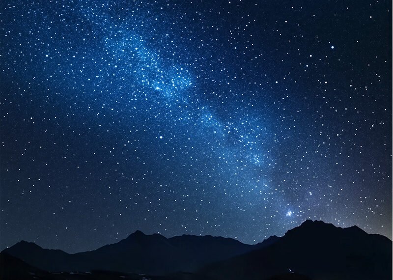 A starry night sky above a silhouette of mountain peaks, with countless stars forming the Milky Way visible across the horizon.