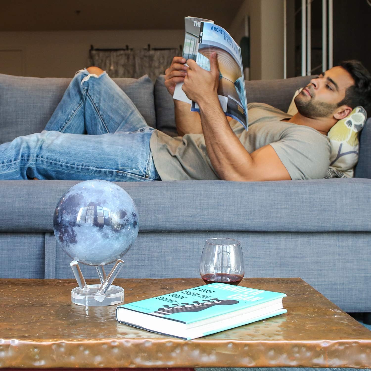 A man in a gray shirt and jeans relaxes on a gray couch reading a magazine. In the foreground, there is a table with a globe, a book, and an empty glass.