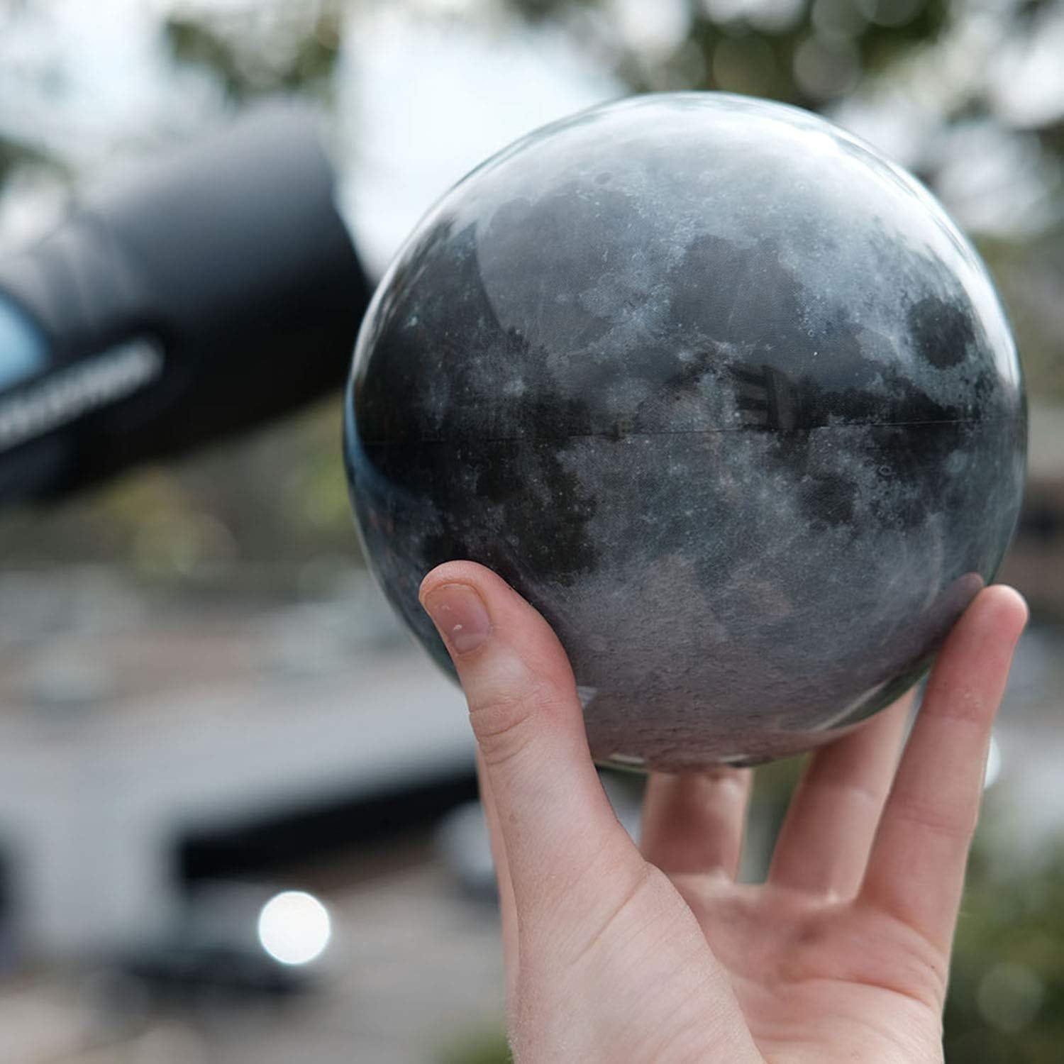 A hand holds a reflective sphere with a moon-like surface while a telescope is visible in the background.