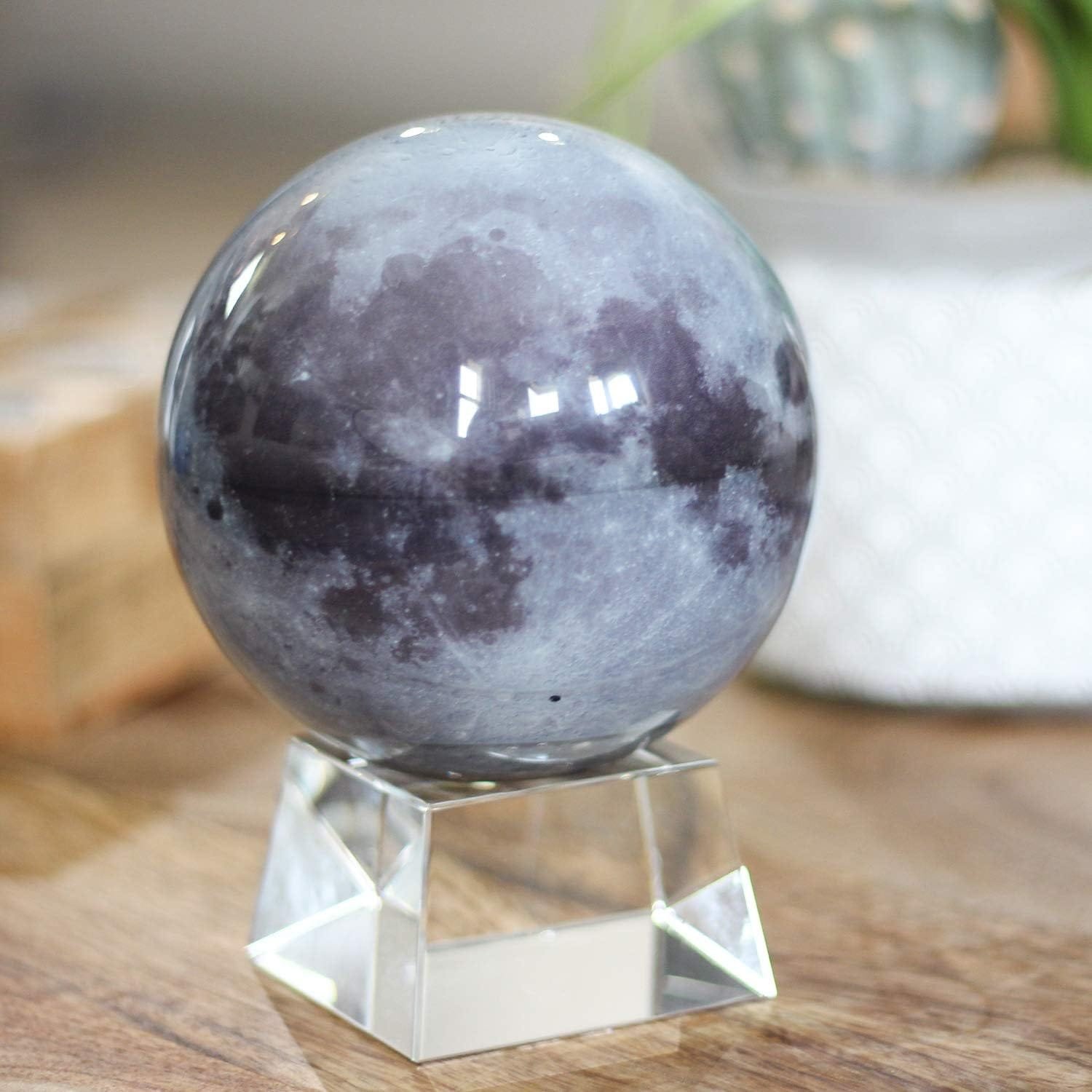 A spherical object with a lunar surface design rests on a transparent square base, placed on a wooden surface; blurred background containing a potted plant.