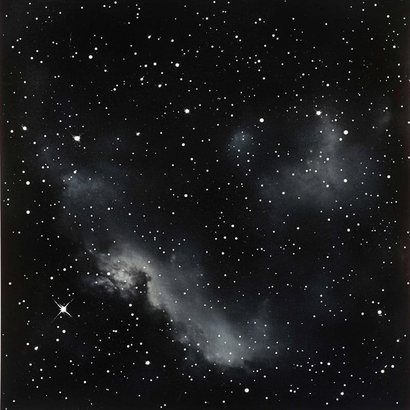 Black and white image of a starry night sky with scattered bright stars and a faint nebula cloud.