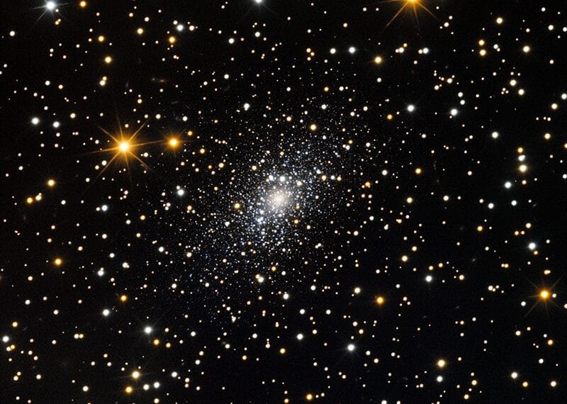 A bright star cluster, shimmering against the black space background, is surrounded by various stars, including the smallest star in the universe.