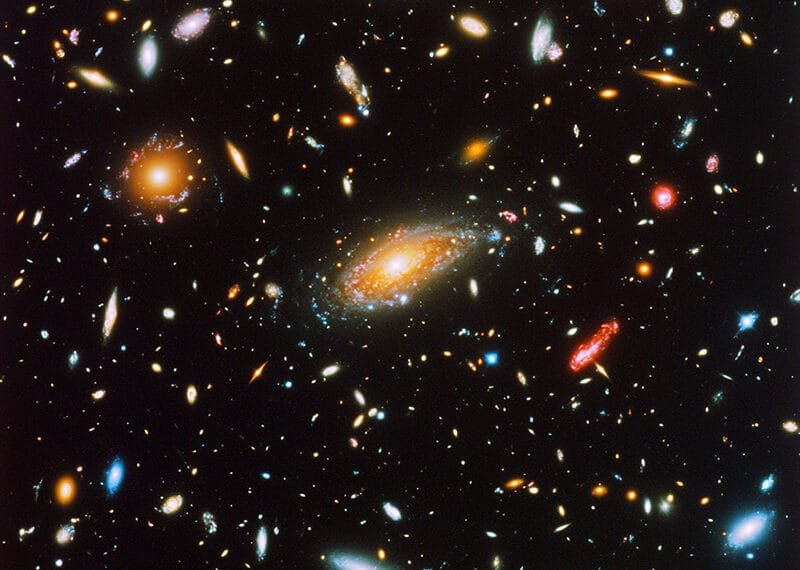 An image of a dense star field with various galaxies in different shapes and colors scattered across the dark galaxy sky.