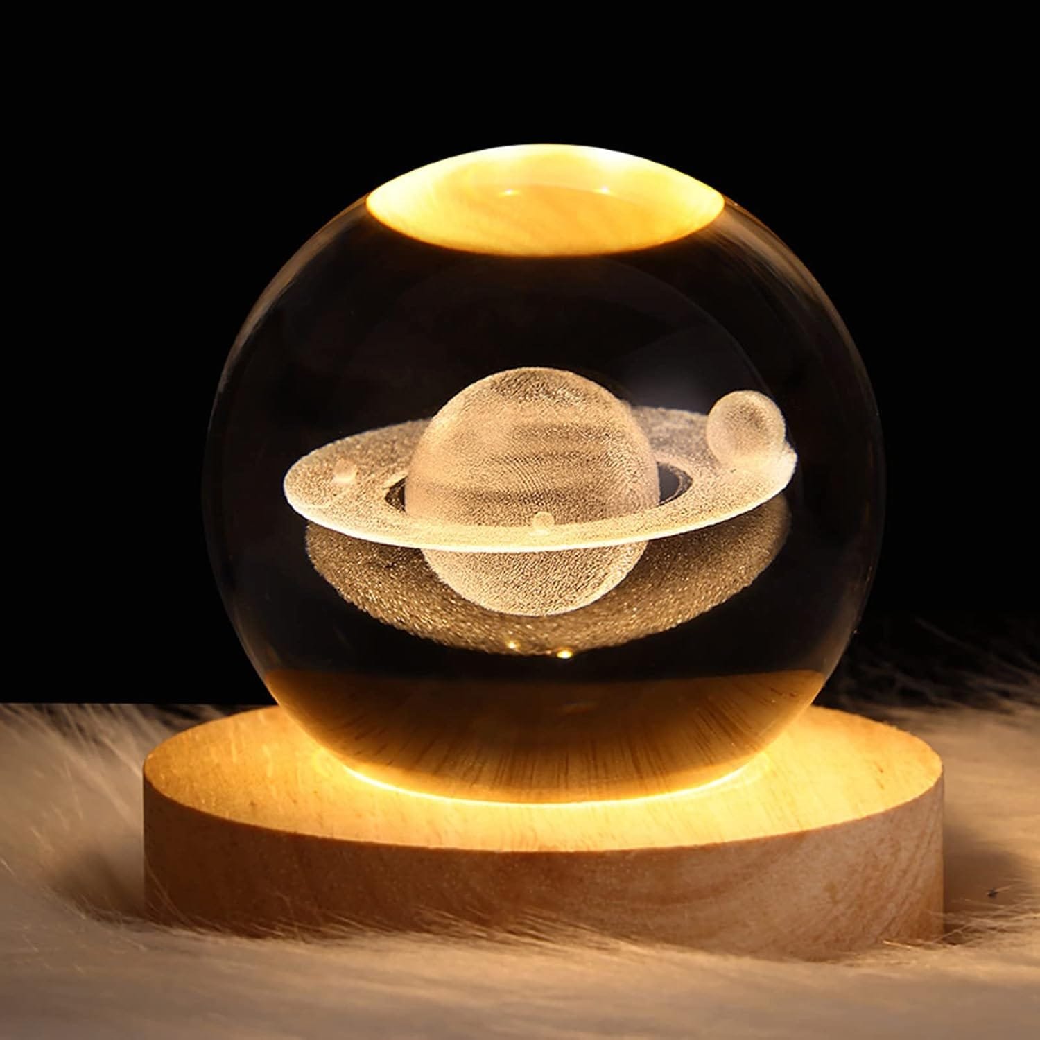 Glass orb with Saturn and rings etched inside, placed on a lit wooden base, against a dark background.