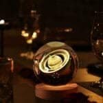 A glowing crystal ball with an image of Saturn sits on a wooden base, surrounded by dimly lit glassware.