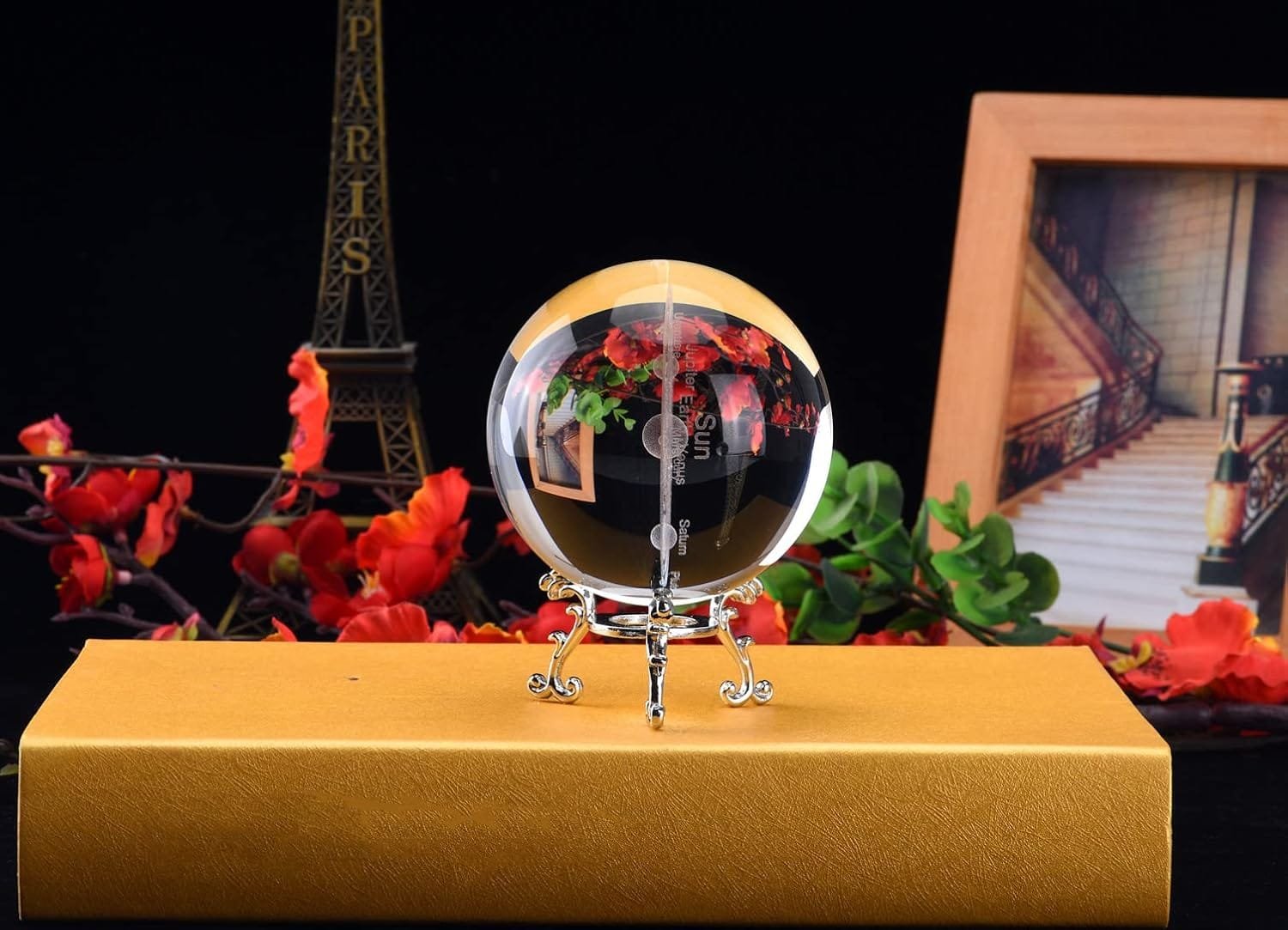 Glass orb with floral design on a silver stand, placed on a gold box. Eiffel Tower model and framed staircase photo in the background.