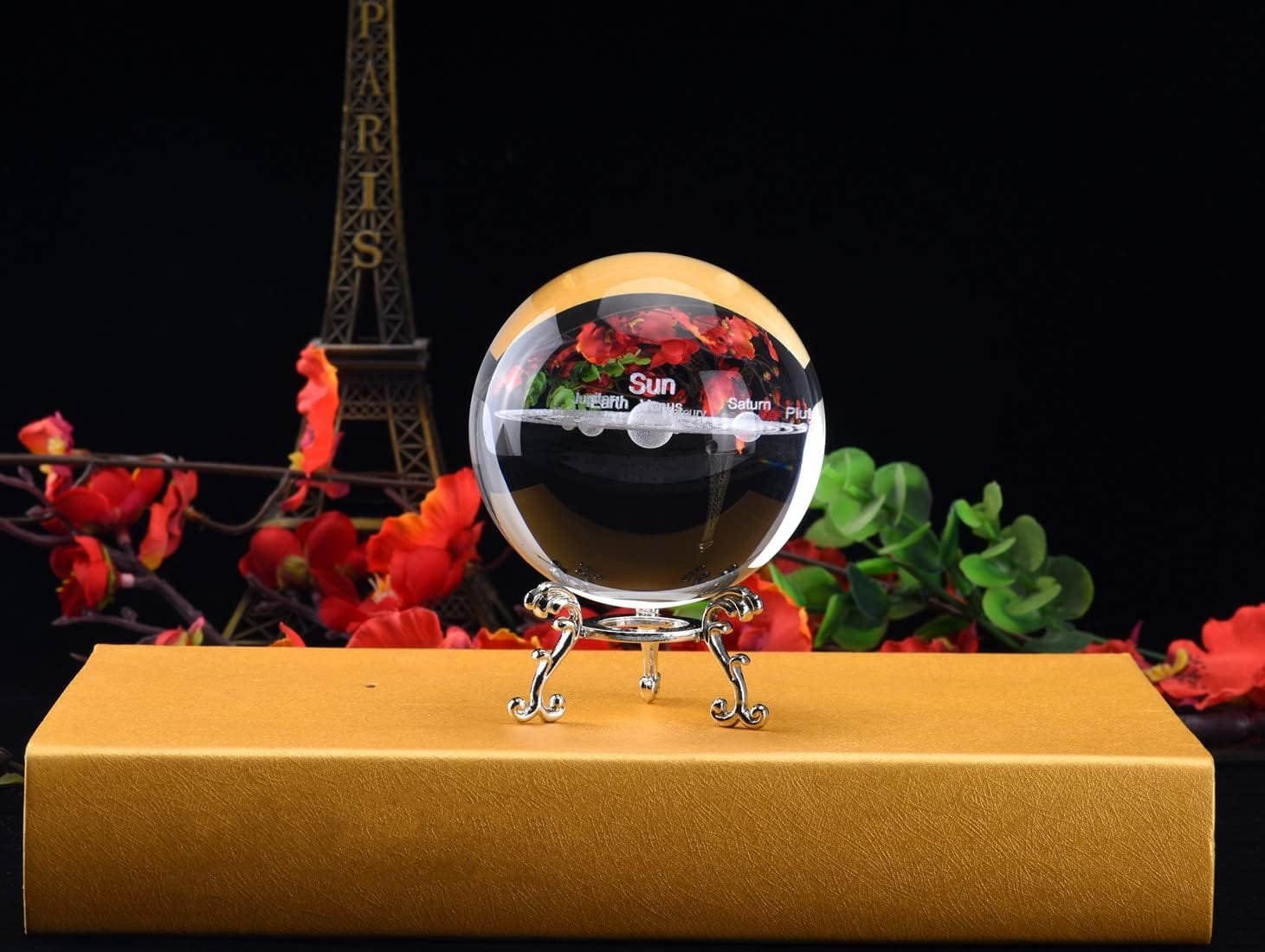 Glass sphere model of the solar system on a yellow box, with red flowers and a miniature Eiffel Tower in the background.