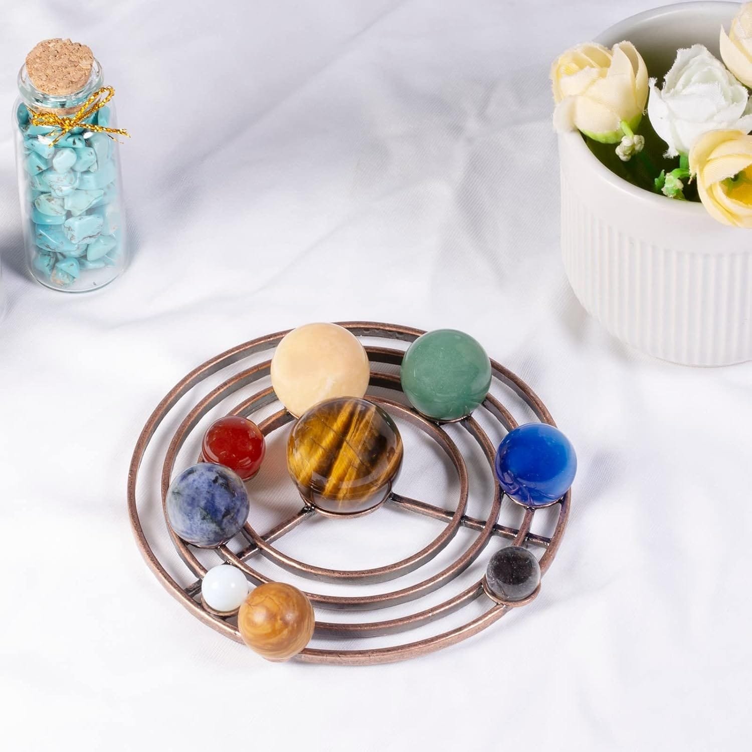 Nine colored stone spheres arranged in a planetary model on a metal framework, with a small jar of turquoise stones and a white pot holding yellow and white flowers in the background.