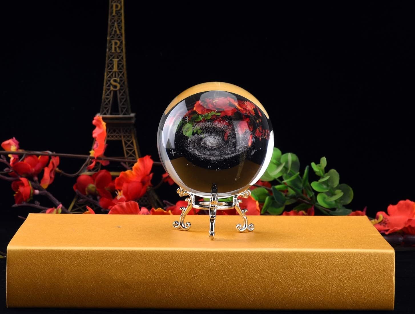 A glass ball displaying a galaxy pattern rests on an ornate stand atop a golden box, with red flowers and a model of the Eiffel Tower in the background.