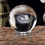 A glass orb with a spiral galaxy design inside, placed on an ornate metal stand. Eiffel Tower model in the background.