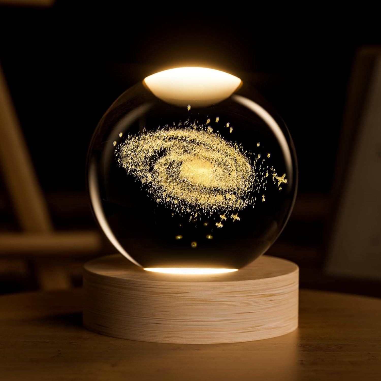 A glowing, spherical lamp displays a spiral galaxy design with small lights resembling stars, resting on a wooden base against a dark background.