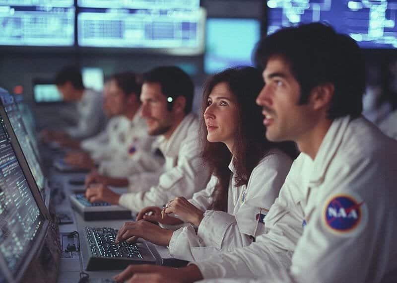 NASA engineers work at computer stations in a control room, with monitors displaying data in the background.