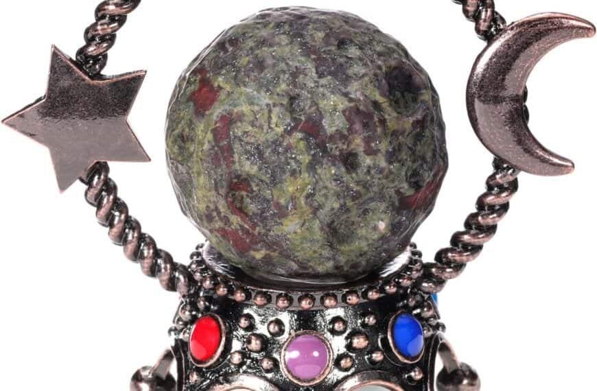 The Jovivi 40mm Natural Crystal Sphere Dragon Blood Jasper Ball Ornament is adorned with metal star and crescent designs, elegantly positioned on a stand embellished with striking red, blue, and purple accents.