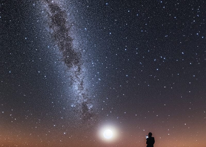 Silhouette of a person standing under a starry night sky, with the Milky Way and a bright moon casting their glow. A lone white star twinkles above the horizon, adding to the celestial wonder.