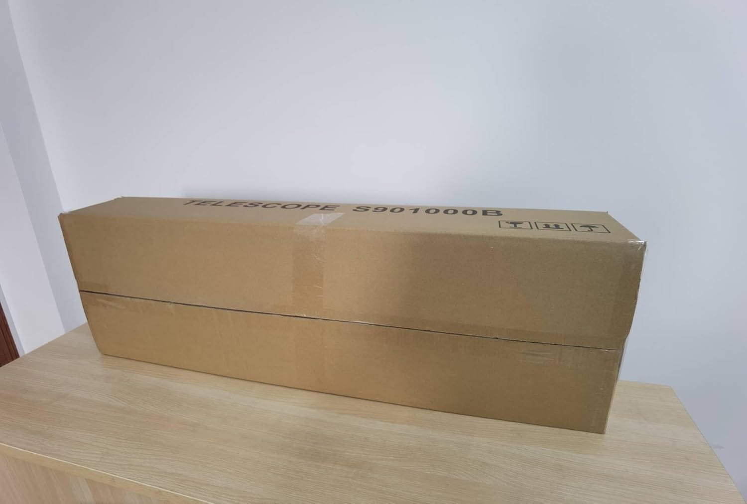 A large brown cardboard box labeled "Telescope S901000B" is placed on a wooden surface against a plain white wall.